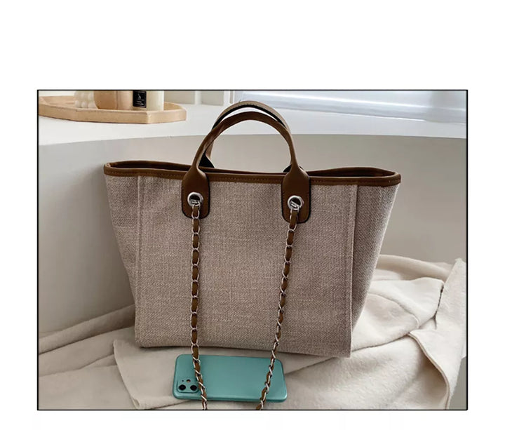 Beige Damenhandtasche aus Leinen mit braunen Ledergriffen und Kettendetails, neben Smartphone.