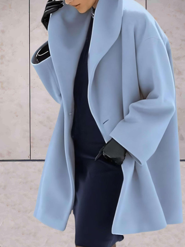 Frau in hellblauem Wollmantel, schwarzer Kleidung und Handschuhen vor beigem Hintergrund.