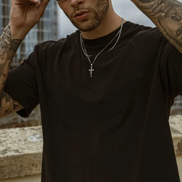 Mann mit schwarzem T-Shirt, Edelstahlkette mit Kreuzanhänger, urbaner Hintergrund.