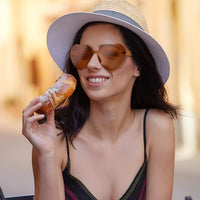 Frau mit Herz-Sonnenbrille und Strohhut genießt Croissant im Freien.