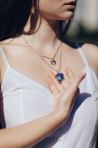 Frau mit blauem Herzanhänger-Halskette, goldene Kette, weißes Top, modisches Accessoire.