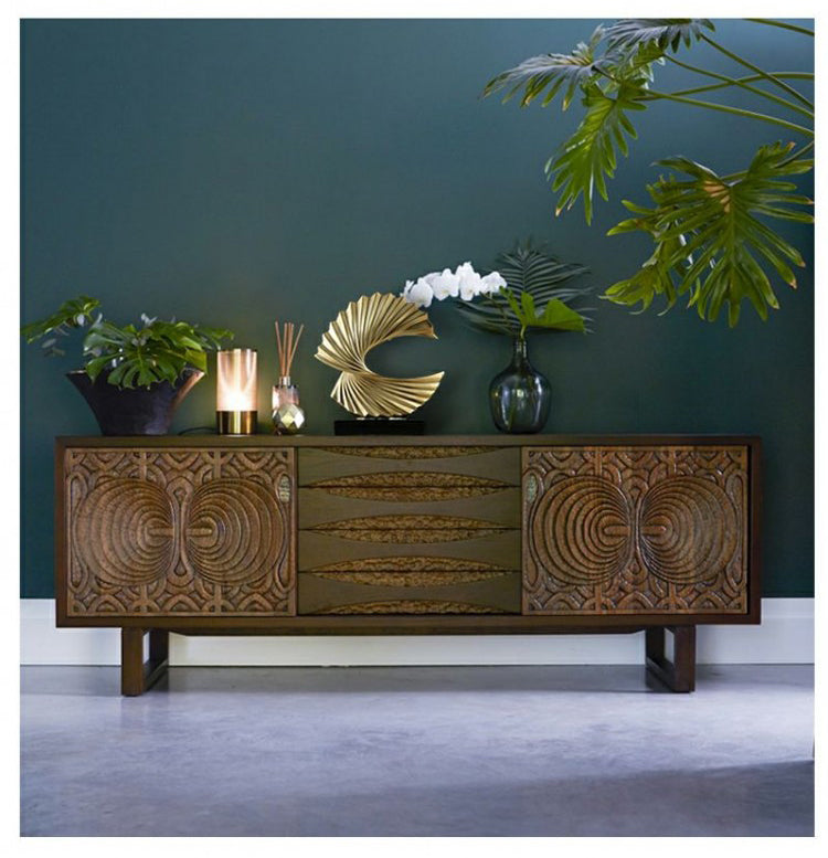 Holz-Sideboard mit geschnitzten Türen, dekoriert mit Pflanzen und Kerzen, vor grüner Wand.