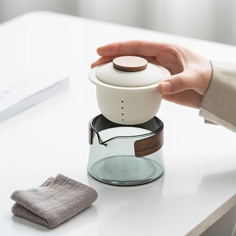 Hand hält weißen Keramik-Teefilter über Glas-Teekanne auf Tisch, modernes Design.