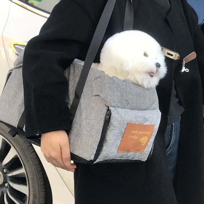 Kleine weiße Hund in grauer Tragetasche mit braunem Lederetikett, getragen von Person in schwarzem Mantel.