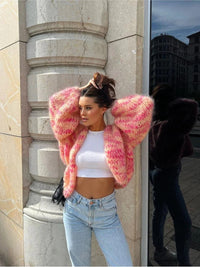 Frau in rosa Strickjacke, weißem Top und Jeans vor Gebäude, modisch, urbaner Stil.