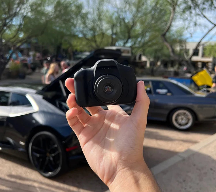 Hand hält schwarze Dashcam vor geparkten Autos im Freien, ideal für Autokamera-Aufnahmen.