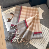 Beigefarbener Karoschal aus Wolle mit Fransen, auf grauem Sofa liegend, neben Zeitschriften.