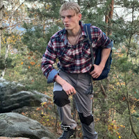 Mann in kariertem Hemd und grauer Hose wandert im Wald, trägt blauen Rucksack.