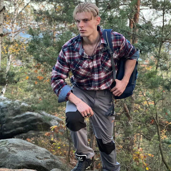 Mann in kariertem Hemd und grauer Hose wandert im Wald, trägt blauen Rucksack.