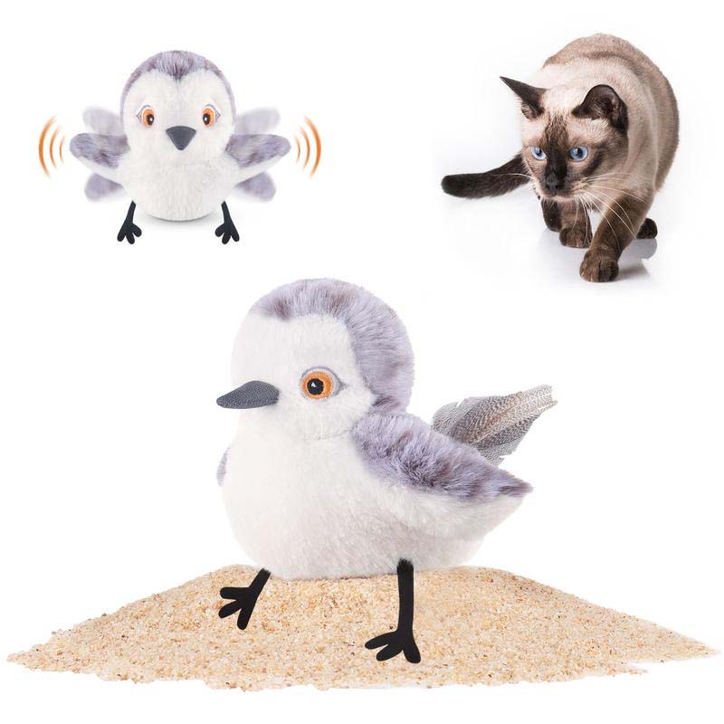 Plüschvogel mit beweglichen Flügeln auf Sand, beobachtet von einer neugierigen Katze.