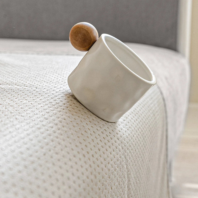 Weiße Keramiktasse mit Holzgriff auf strukturiertem, grauem Stoffhintergrund.