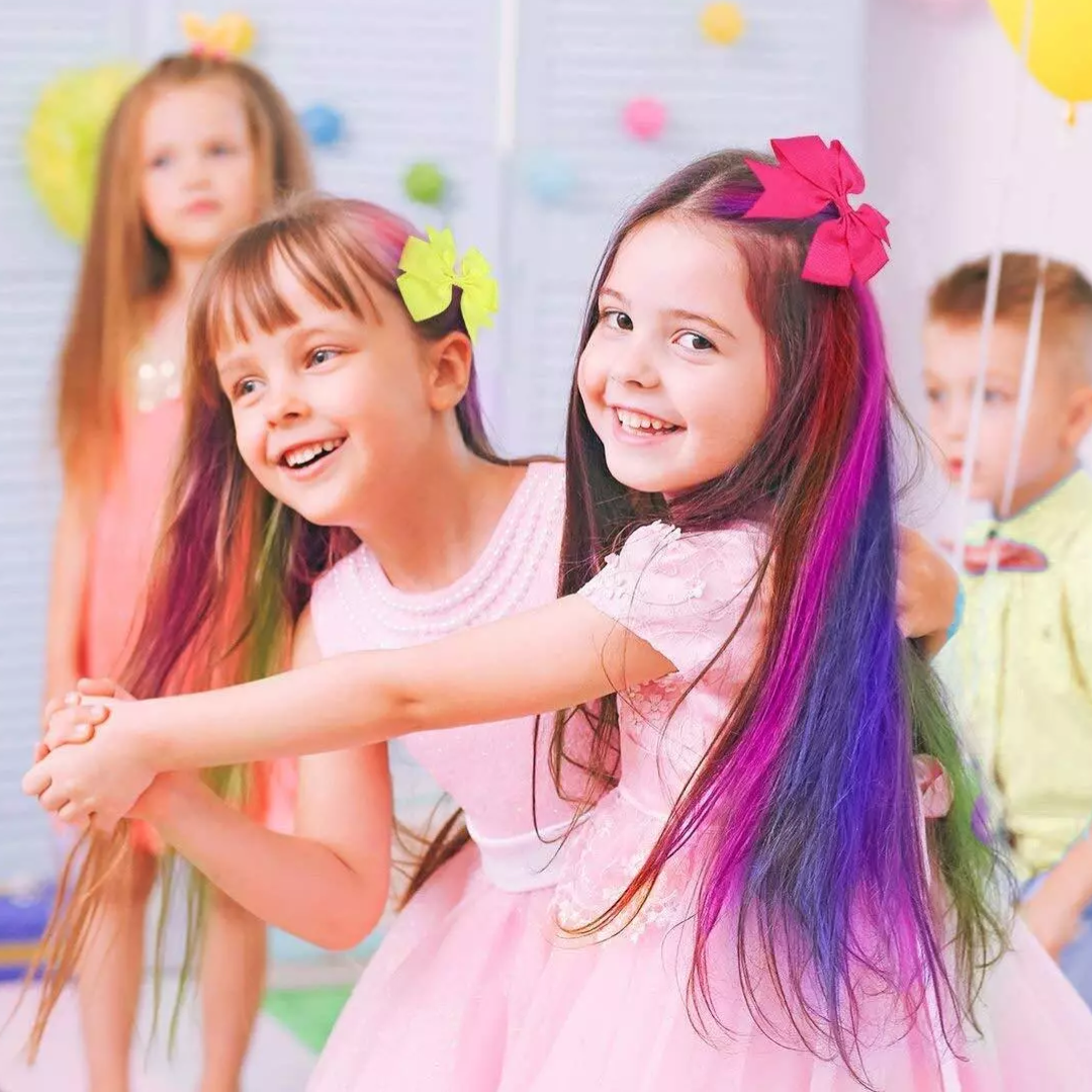 Zwei Mädchen mit bunten Haaren und Schleifen tanzen fröhlich in einem pastellfarbenen Raum.