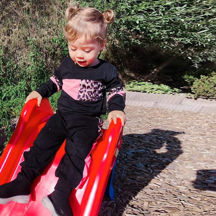 Kleinkind in schwarzem Outfit mit Leopardenmuster rutscht auf roter Rutsche im Freien.