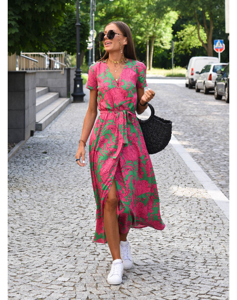 Frau im pink-grünen Sommerkleid, trägt schwarze Tasche, weiße Sneaker, Sonnenbrille, lächelnd.