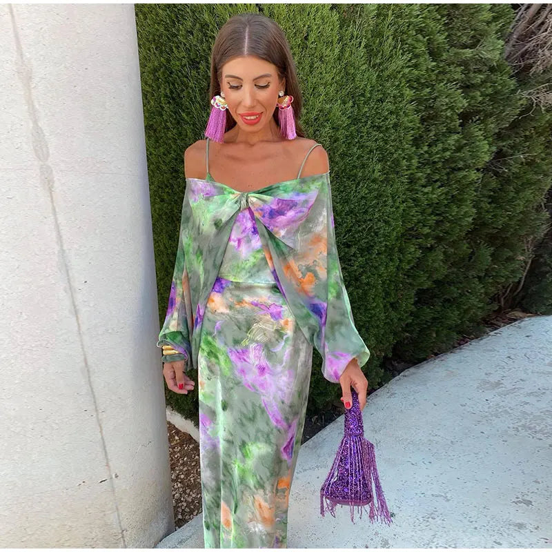 Frau in grün-lila Kleid mit Fransenohrringen und lila Tasche vor grüner Hecke.