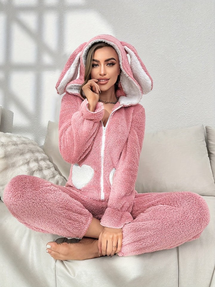 Frau in rosa Plüsch-Hasen-Einteiler mit Kapuze, sitzt auf Sofa, gemütliche Loungewear.