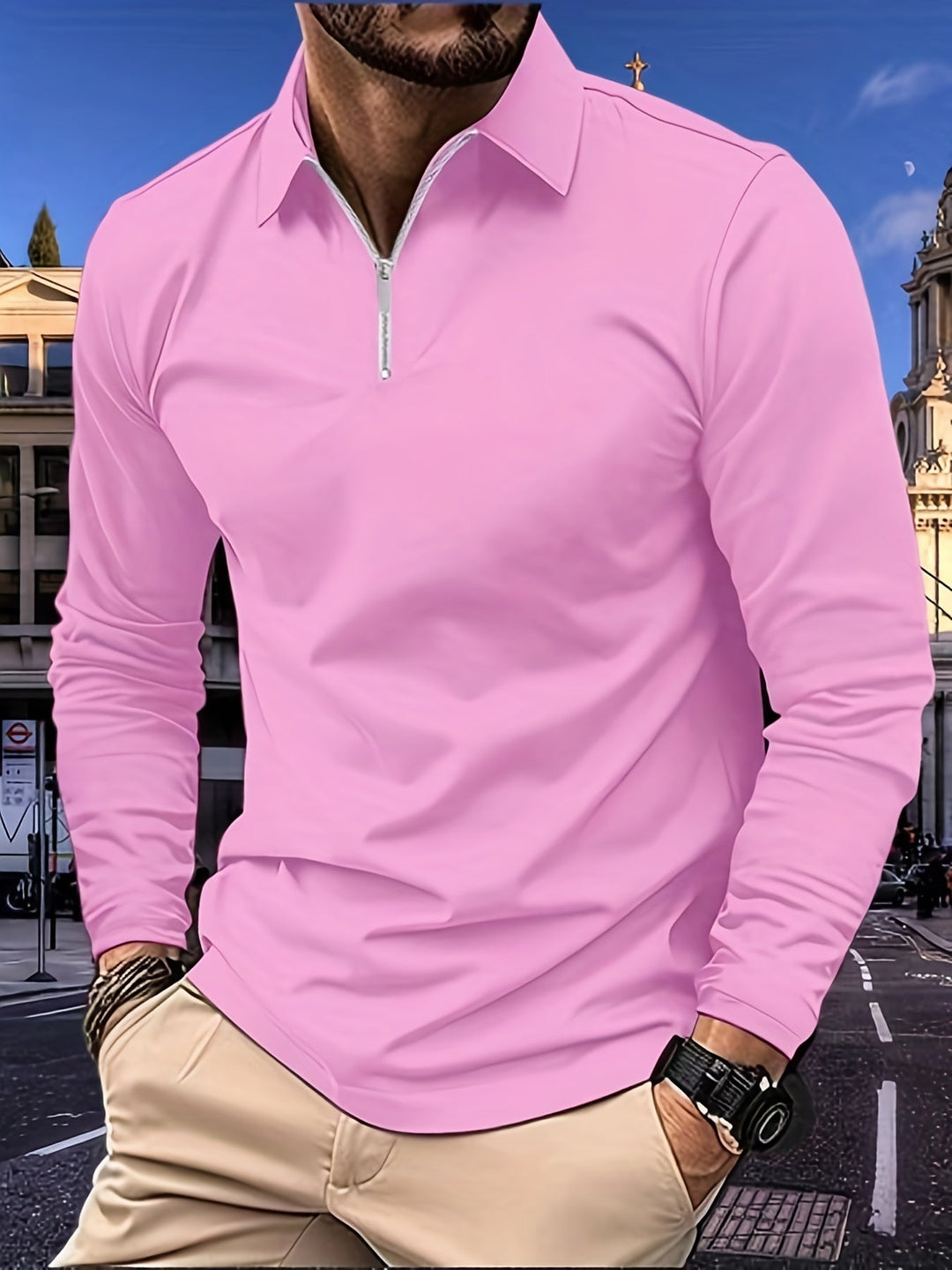 Mann in rosa Langarmshirt mit Reißverschluss, beige Hose, Stadt im Hintergrund.