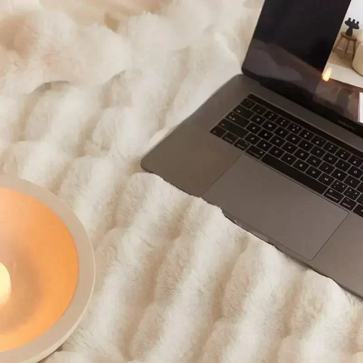 Laptop auf weißer, flauschiger Decke neben Kerze, gemütliches Home-Office-Setup.