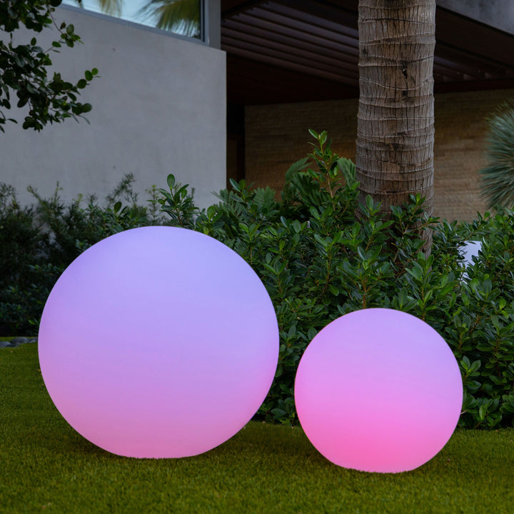 Zwei leuchtende LED-Gartenkugeln, rosa und lila, auf Rasen vor modernem Haus.