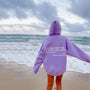 Frau am Strand in lila Hoodie mit Aufschrift, Meer im Hintergrund, bewölkter Himmel.