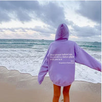 Person in lila Hoodie am Strand, Ozean im Hintergrund, inspirierender Text auf dem Rücken.