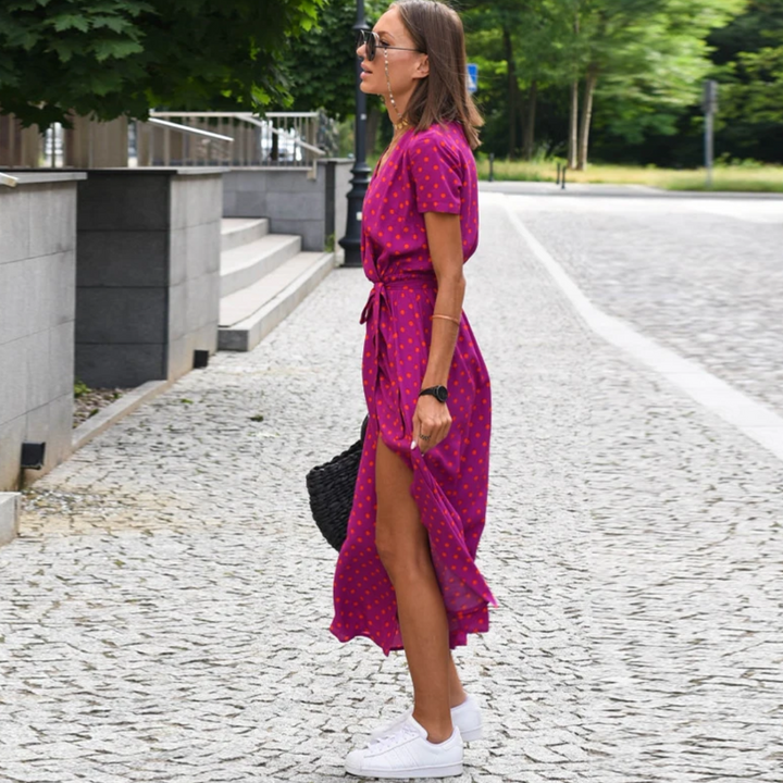 Frau in pinkem Sommerkleid mit Punkten, weiße Sneaker, schwarze Tasche, auf Kopfsteinpflaster.