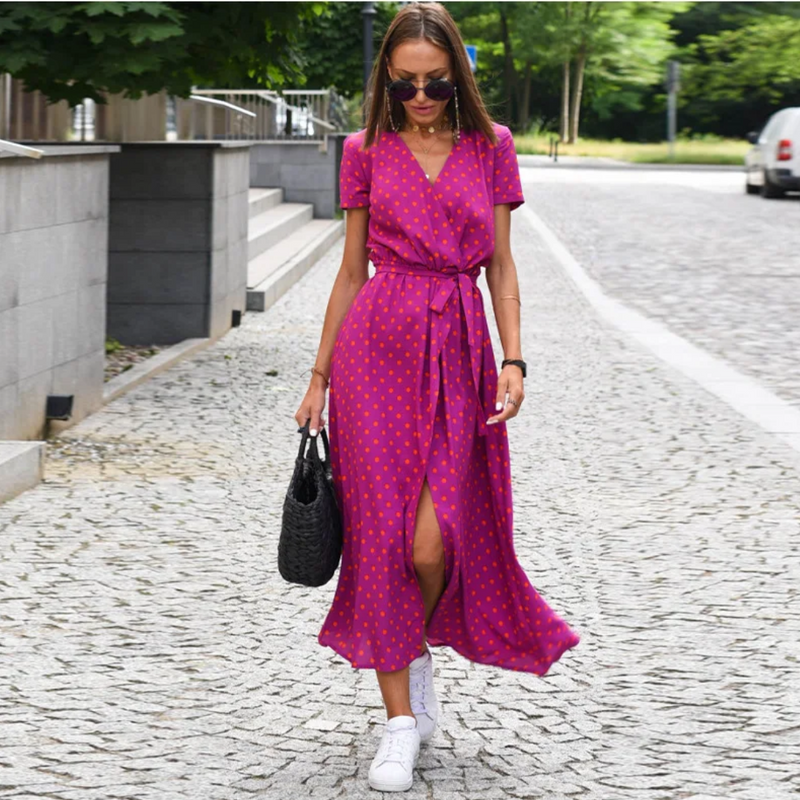 Frau im pinken Sommerkleid mit Punkten, trägt schwarze Tasche und weiße Sneaker auf Kopfsteinpflaster.