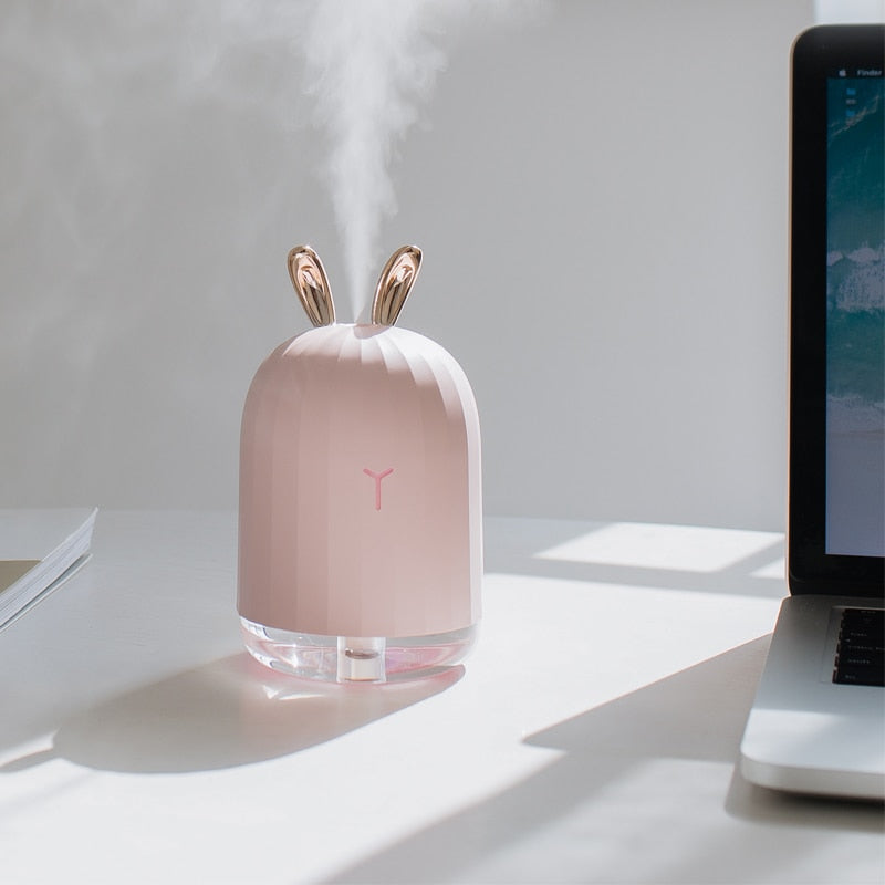 Rosa Luftbefeuchter mit Hasenohren, USB-betrieben, auf Schreibtisch neben Laptop.