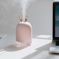 Rosa Luftbefeuchter mit Hasenohren, USB-betrieben, auf Schreibtisch neben Laptop.