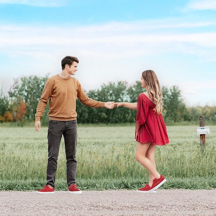 Paar in roter Kleidung hält Hände, steht auf Feldweg vor grünem Feld und blauem Himmel.
