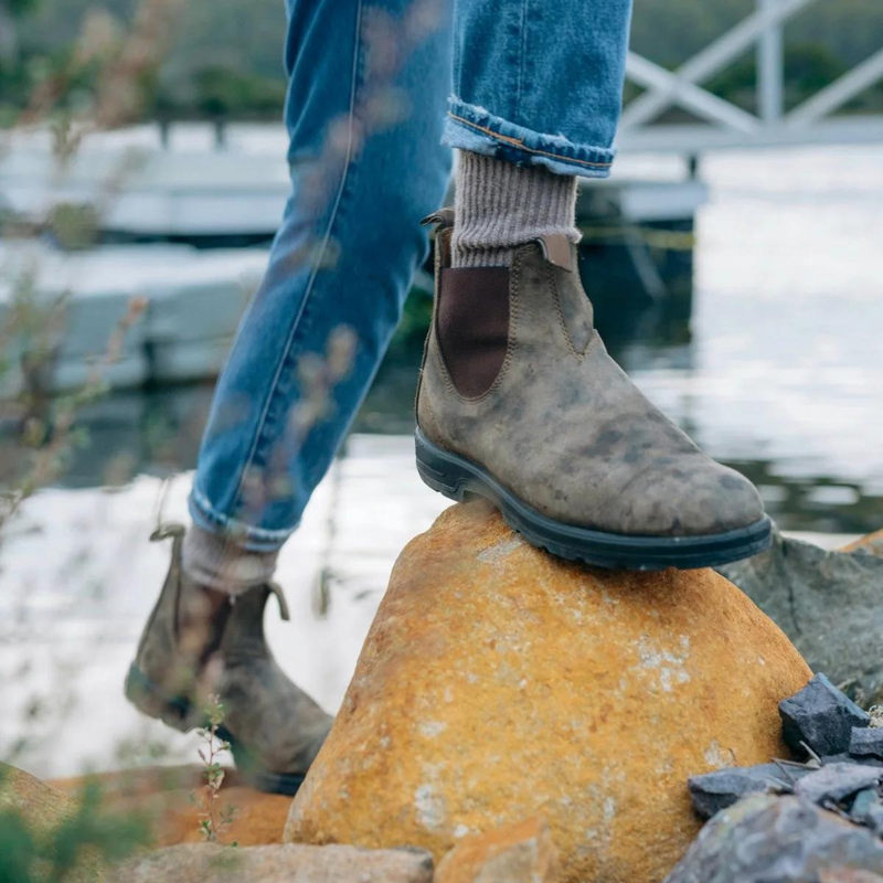 Person trägt braune Lederstiefel auf Felsen am See, Outdoor-Wanderschuhe, Jeans, Natur.
