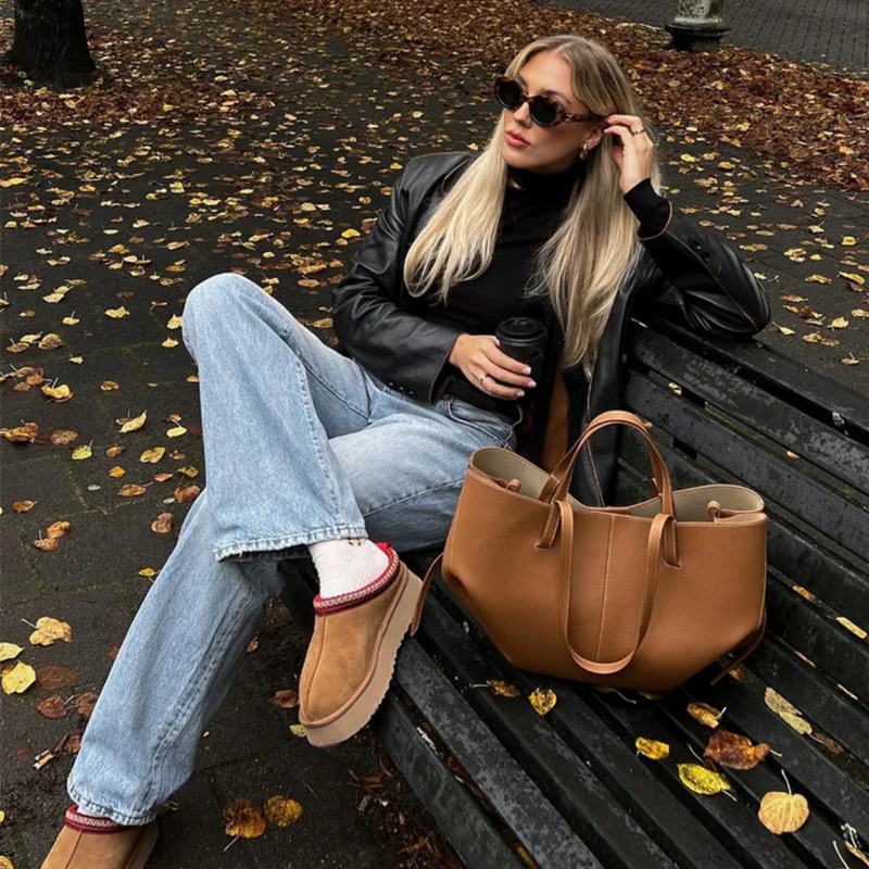 Frau mit brauner Ledertasche auf Parkbank, Herbstmode, Sonnenbrille, lässiger Stil.