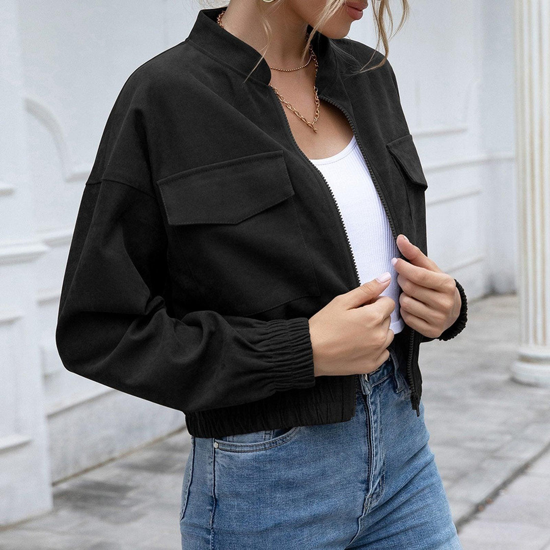 Frau in schwarzer Bomberjacke, weißem Top und blauer Jeans vor weißer Wand.