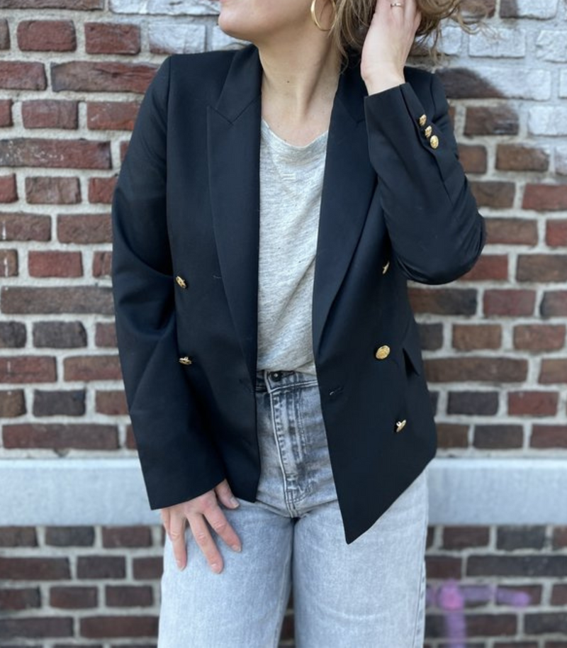 Frau in schwarzem Blazer mit goldenen Knöpfen, grauem T-Shirt und Jeans vor Ziegelwand.