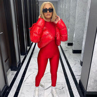 Frau in rotem Jumpsuit und roter Daunenjacke, blonde Haare, schwarze Sonnenbrille, weiße Turnschuhe.