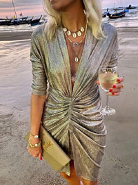 Frau in silbernem Glitzerkleid am Strand, hält Weinglas, trägt goldene Accessoires.