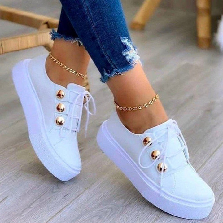 Weiße Damen-Sneaker mit goldenen Knöpfen, Jeans, Holzfußboden, modische Freizeitschuhe.
