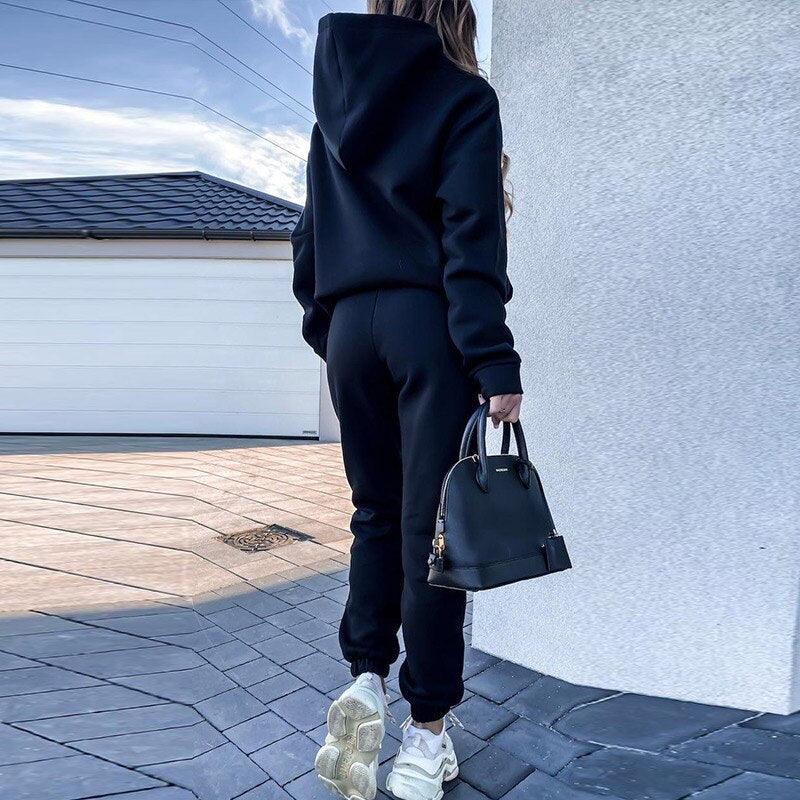 Frau in schwarzem Hoodie und Jogginghose, weiße Sneaker, schwarze Handtasche, urbaner Hintergrund.