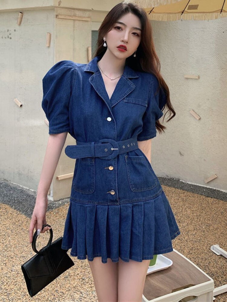 Frau in blauem Jeanskleid mit Puffärmeln und Gürtel, hält schwarze Handtasche.