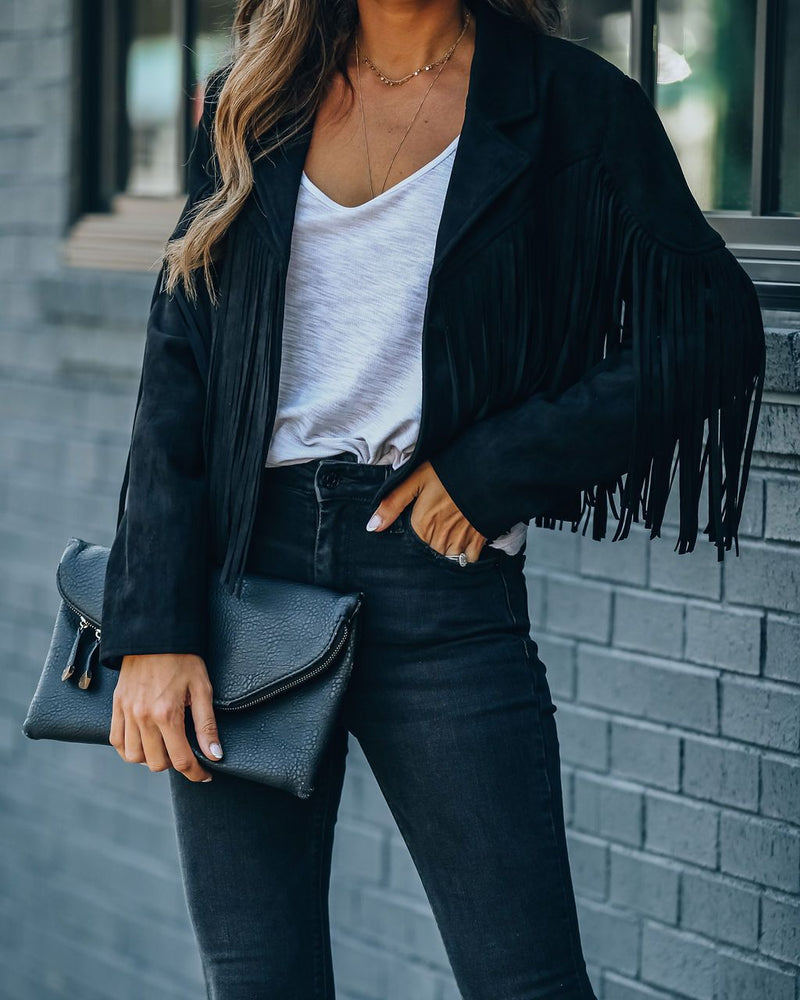 Frau in schwarzer Fransenjacke, weißem Top, schwarzer Jeans, hält schwarze Clutch.