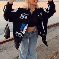 Frau in schwarzer NASCAR-Jacke mit Logos, weite Jeans, Sonnenuntergang am Wasser.