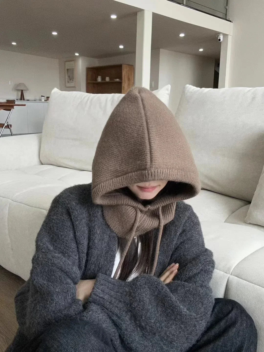 Frau mit braunem Strick-Balaclava und grauem Pullover auf weißem Sofa in modernem Wohnzimmer.