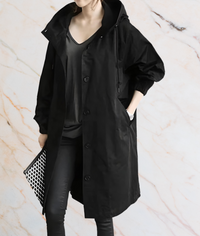 Frau in schwarzem Regenmantel mit Kapuze, hält schwarz-weiße Clutch, modisch, elegant.