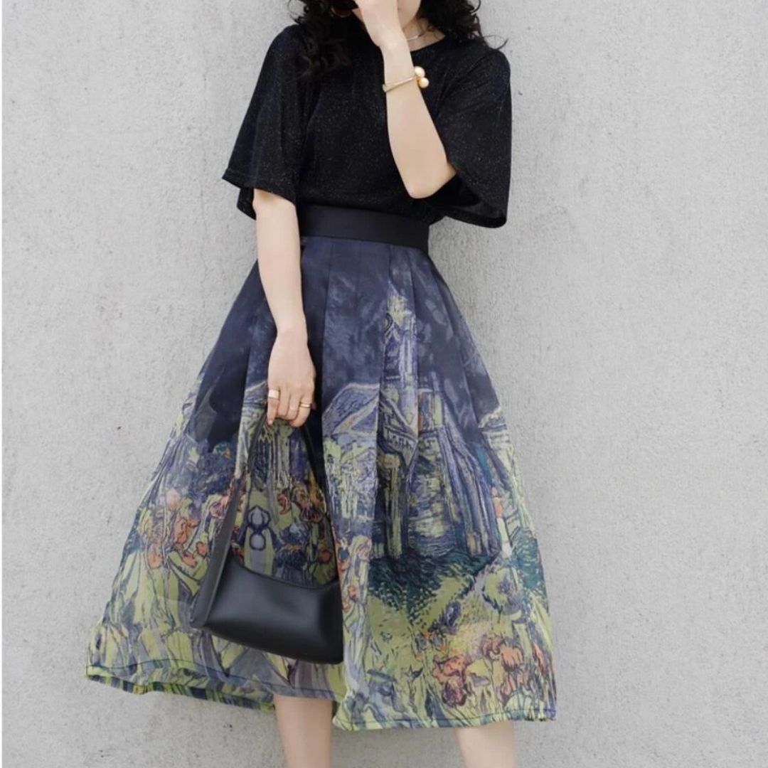 Frau in schwarzem T-Shirt und bedrucktem Rock, hält schwarze Handtasche, vor grauer Wand.