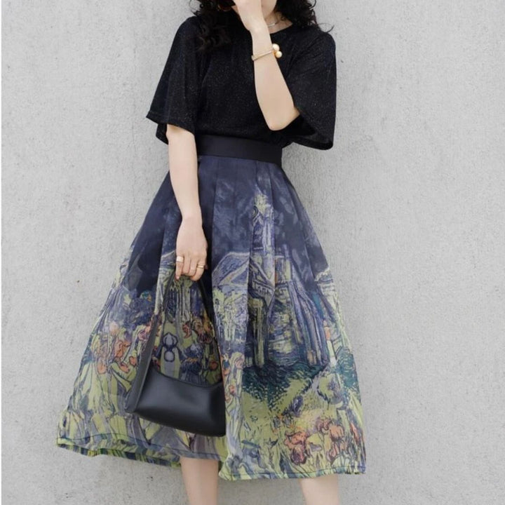 Frau in schwarzem T-Shirt und bedrucktem Rock, hält schwarze Handtasche, vor grauer Wand.