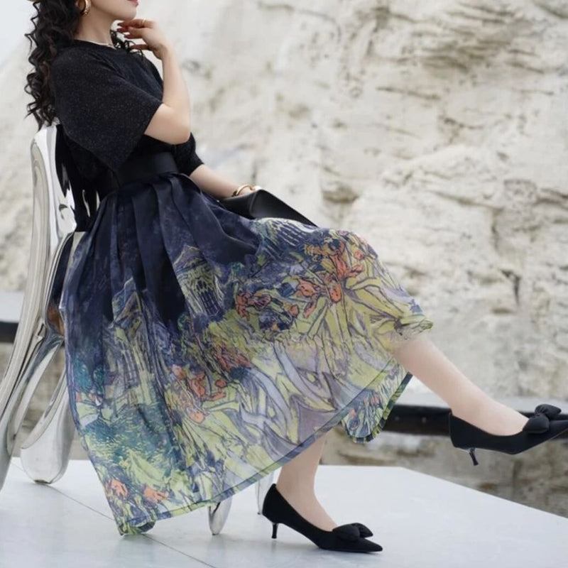 Frau in schwarzem Kleid mit buntem Rock, sitzt auf Stuhl, elegante schwarze Schuhe.