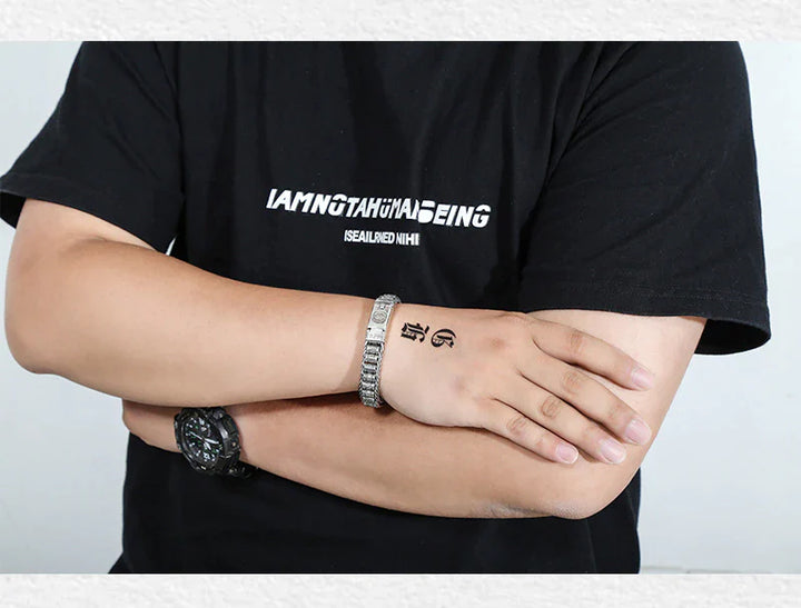 Person mit schwarzem T-Shirt, Armbanduhr und Armband, verschränkte Arme, tätowierte Hand.