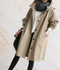 Frau in beigem Trenchcoat, schwarzer Hose, weißem Pullover, grauem Schal vor Marmorhintergrund.