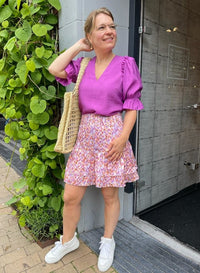 Frau in lila Bluse und bunter Rock, trägt Strohtasche, weiße Sneaker, steht vor grünem Hintergrund.