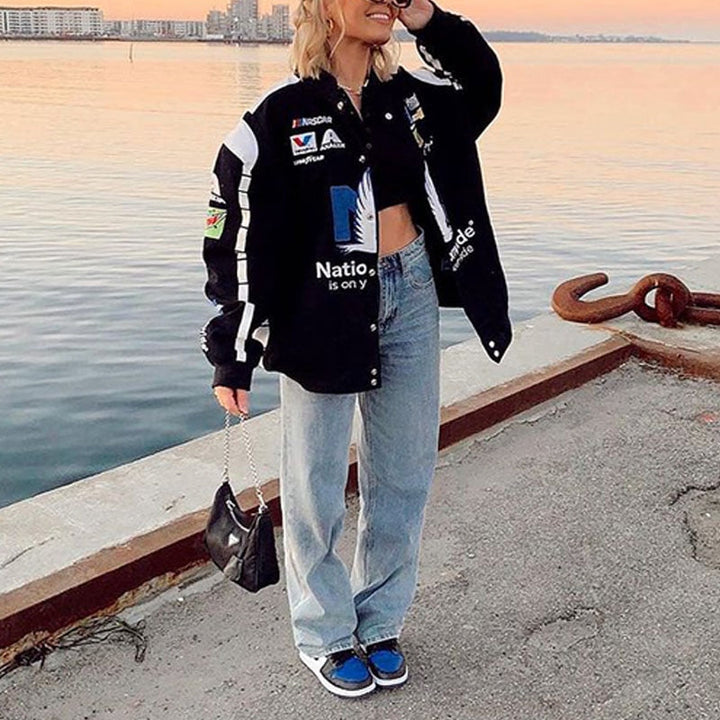 Frau in schwarzer Rennjacke, blauer Jeans und blauen Sneakers am Hafen bei Sonnenuntergang.
